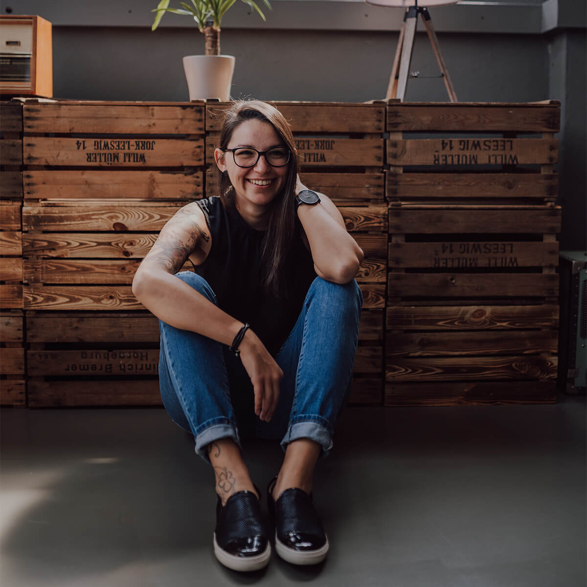 Kerstin Partheder, Design & Illustration, Grafikerin aus Linz, sitzend vor Holzkiste, lachend mit Tatowierung auf rechtem Arm